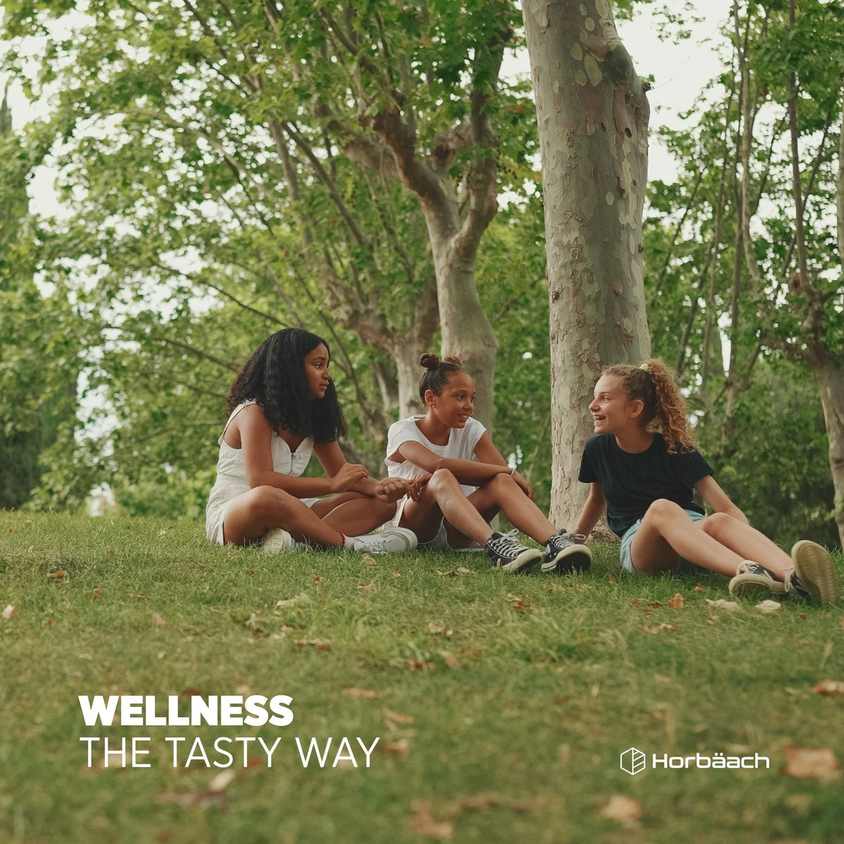 Three young girls sitting on grass and chatting in a park. Text on image: WELLNESS THE TASTY WAY, Horbäach.