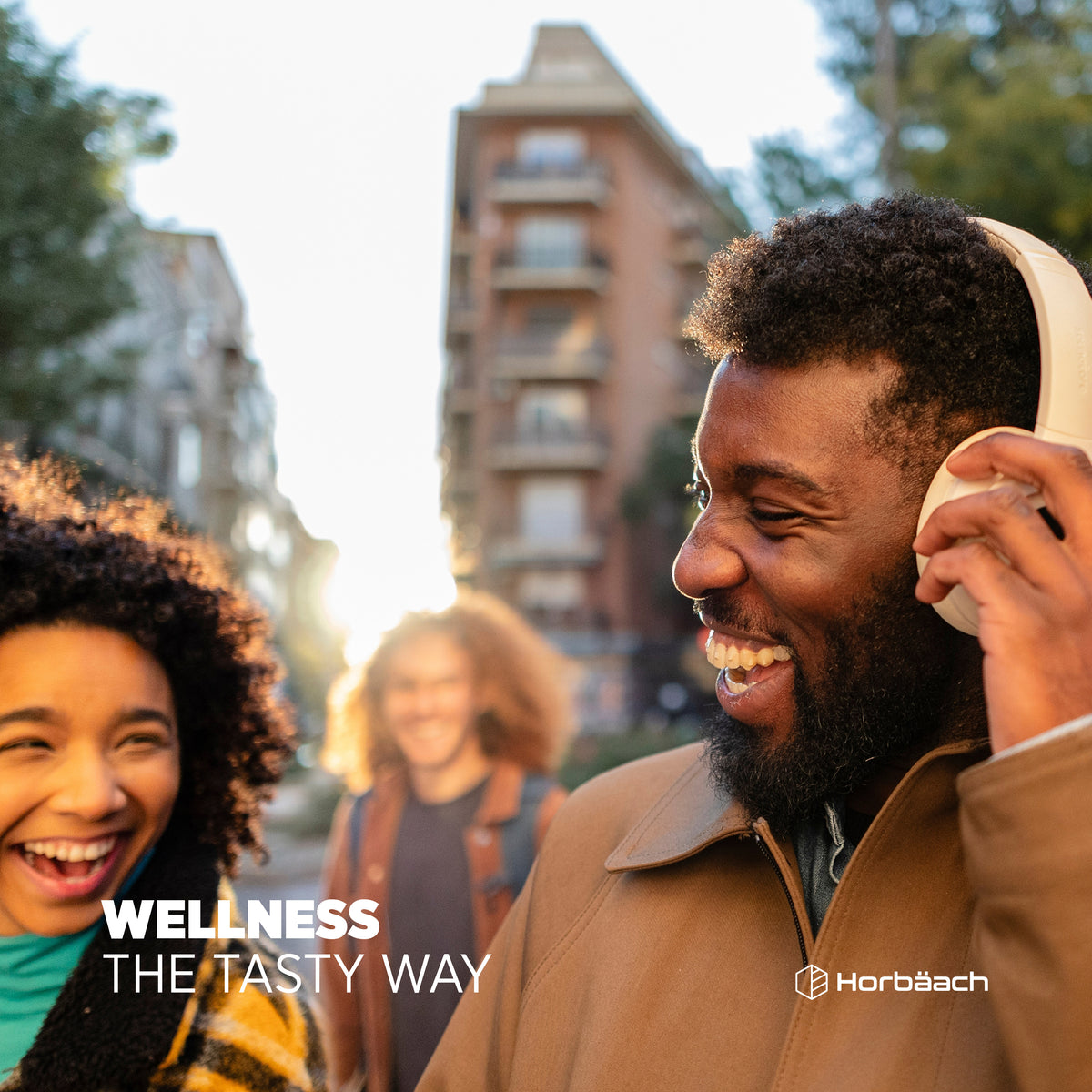 Smiling man with headphones and laughing woman walk outdoors on a sunny day, with another person blurred behind them.