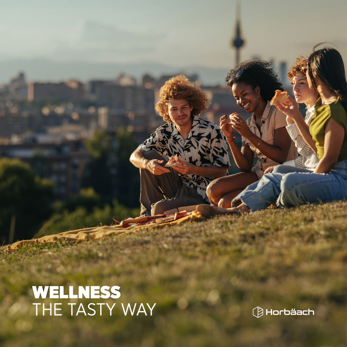 Young adults picnicking on a grassy hill, eating pizza with a city skyline, featuring the text 'Wellness The Tasty Way'.
